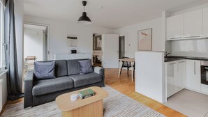 A modern apartment with a grey sofa, a wooden coffee table, a kitchen with white cabinets, and a dining area with a wooden table and chairs.