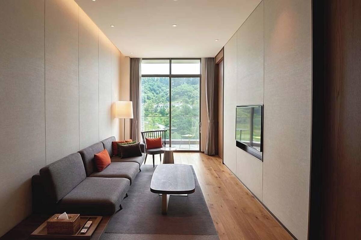 A modern living room with a sofa, a coffee table, a TV mounted on the wall, and a view of greenery through large windows.