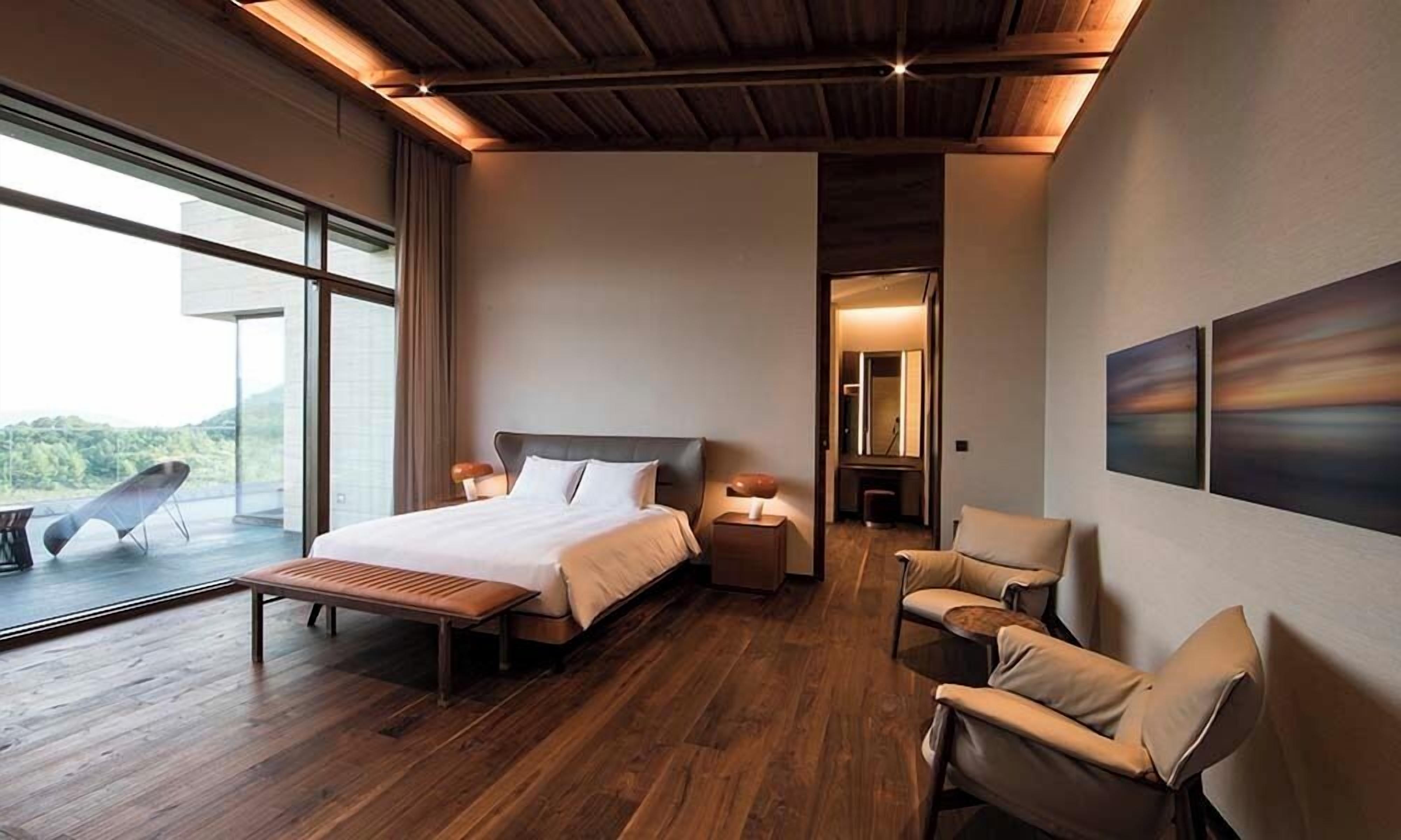 A modern bedroom with a large bed, a bench, two chairs, and a view of the outdoors.