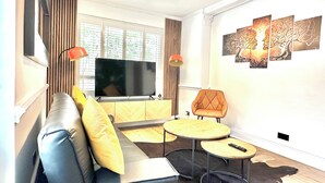 Ein modernes Wohnzimmer mit einem Ledersofa, einem Flachbildfernseher, zwei kleinen Holztischen und einem großen Fenster mit Jalousien.