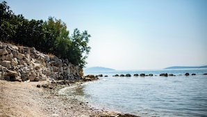 Eine felsige Küste mit einer Steinmauer und Bäumen, die zu ruhigem Wasser und fernen Bergen führt.