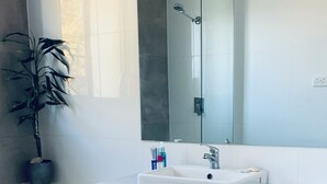 Une salle de bain moderne avec un lavabo blanc, un grand miroir, une douche et une plante en pot.