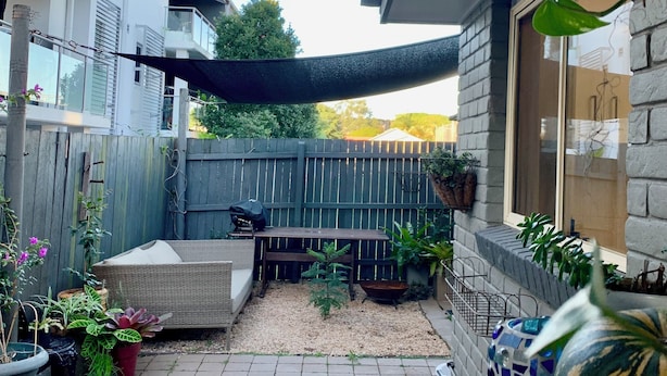 Un patio couvert avec une table et des chaises en bois, entouré de plantes en pot et d’un barbecue.
