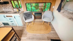 Un intérieur chaleureux avec un coin cuisine, deux fauteuils dotés de coussins moelleux, une cheminée et une vue sur la verdure par une fenêtre.