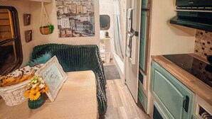 Un espace de vie compact comprenant un coin cuisine, un lit agrémenté d’une tête de lit décorative, un téléviseur fixé au mur et une fresque représentant un paysage urbain.