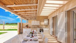 An outdoor dining area with a table set for four, white chairs, a kitchen with white cabinets, and a view of the countryside.