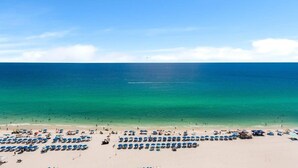 Ein Strand mit Reihen aus blauen Sonnenschirmen und Liegestühlen, einem klaren Himmel und dem Meer im Hintergrund.
