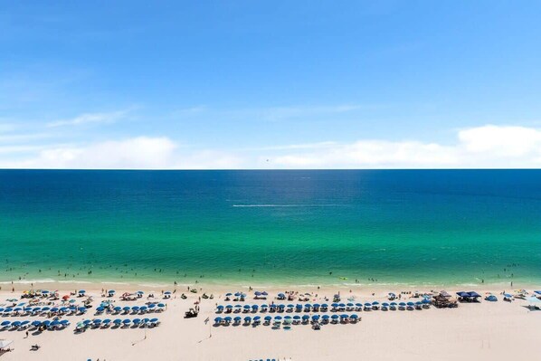 Ein Strand mit Reihen aus blauen Sonnenschirmen und Liegestühlen, einem klaren Himmel und dem Meer im Hintergrund.