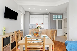 A modern dining area with a table set for four, wooden chairs, a TV mounted on the wall, and a kitchen in the background.
