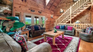 A cozy log cabin interior with a wooden staircase, two sofas, a coffee table, and a large window overlooking greenery.