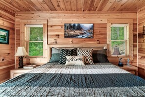 A wooden cabin bedroom with a bed, a painting of a forest scene, and two windows with blinds.