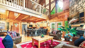A spacious wooden cabin with a dining area, kitchen, and a cozy living room with a red rug and multiple seating options.
