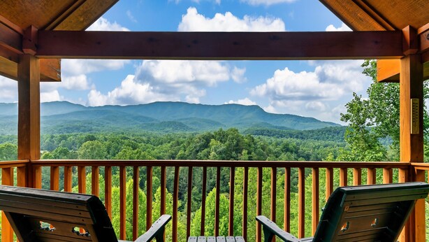 A wooden deck with two chairs and a table, overlooking a forested mountain range.
