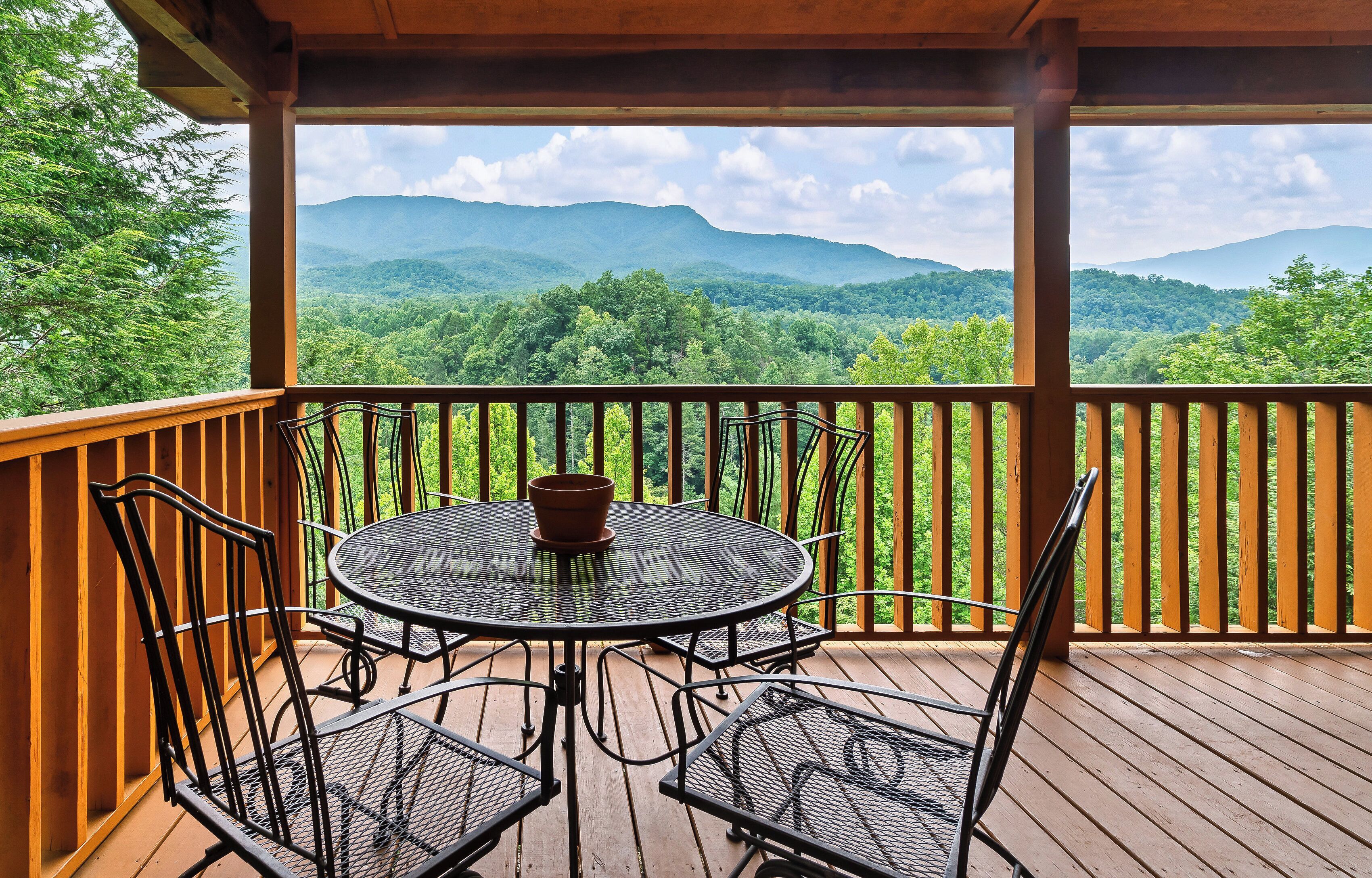 A wooden deck with a round table and four chairs, overlooking a forested mountain range.