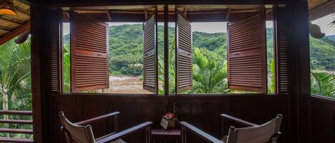 A room with two wooden chairs facing a window with open shutters, overlooking a lush green landscape.