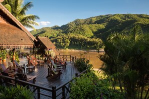 A wooden deck with outdoor seating, overlooking a river and lush greenery. - The Luang Say Lodge (Pakbeng)