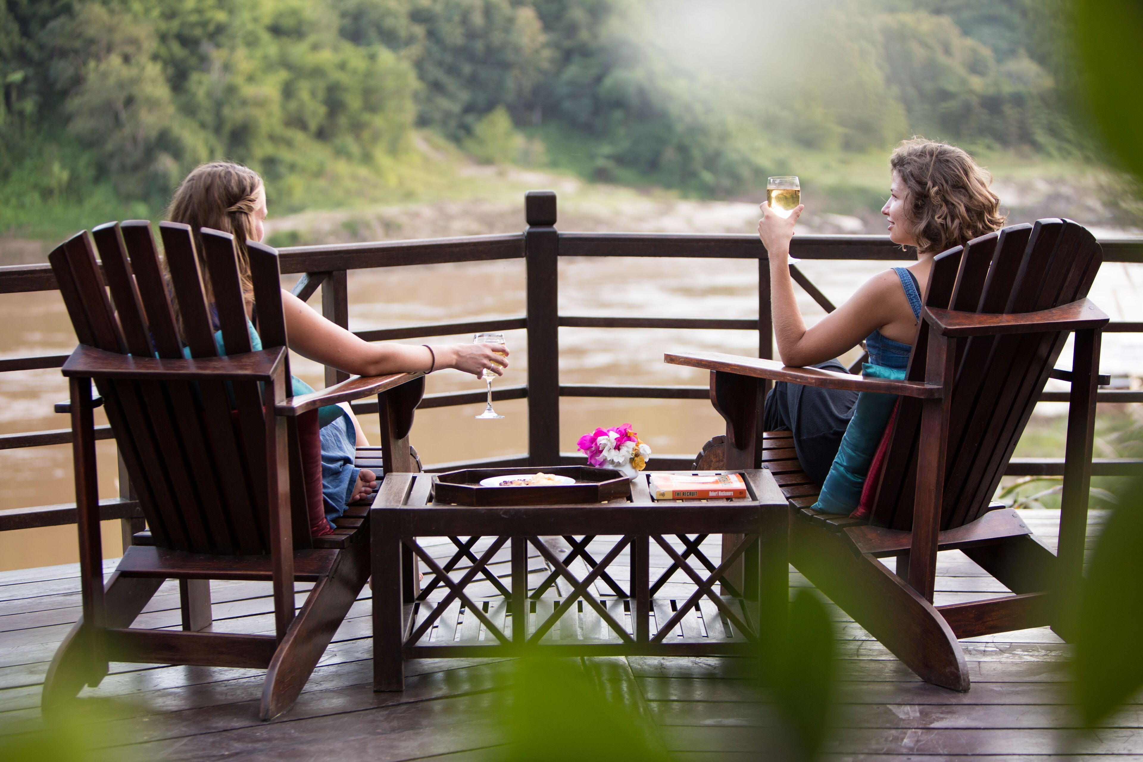 Two people seated on wooden chairs at a table on a wooden deck with a river view.
