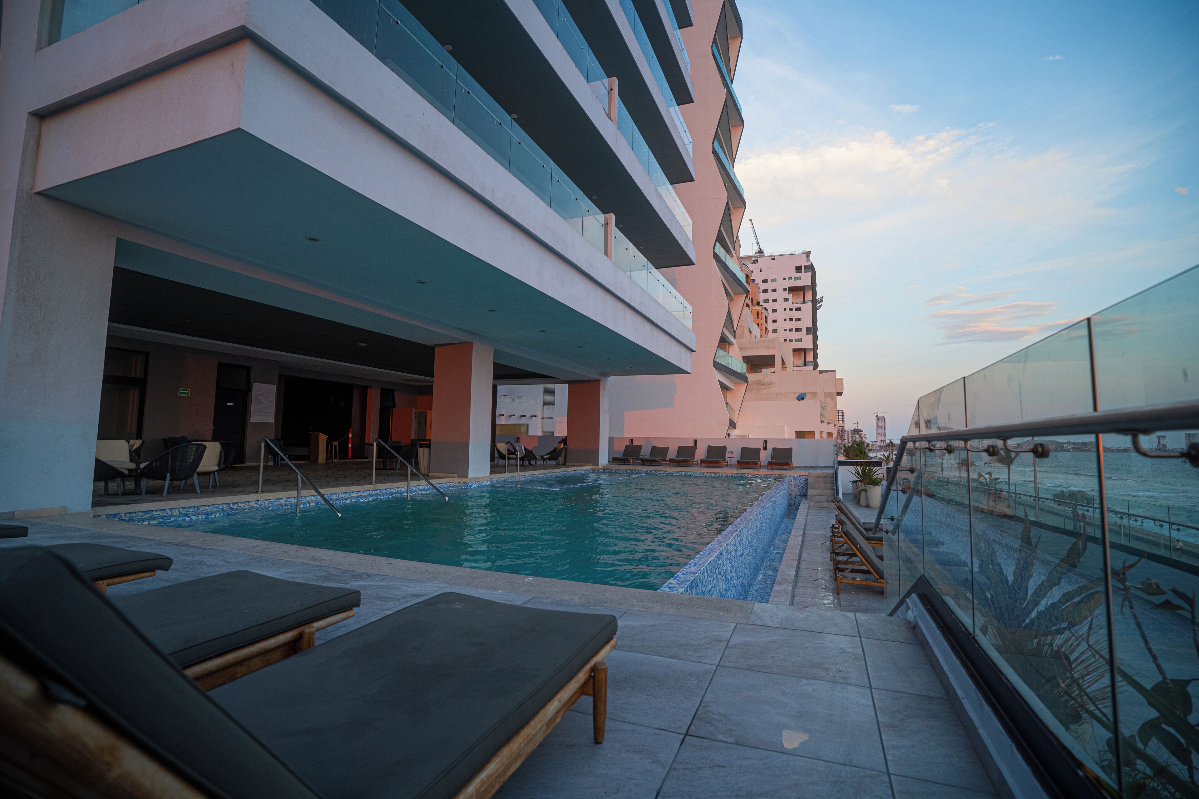 A rooftop pool area with lounge chairs, a pool, and a glass railing overlooking the city.