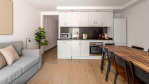 Une cuisine moderne avec des armoires blanches, un four à micro-ondes, une cuisinière et un réfrigérateur. On y trouve une table à manger en bois avec des chaises. Un canapé gris et une plante se trouvent dans l’espace salon.