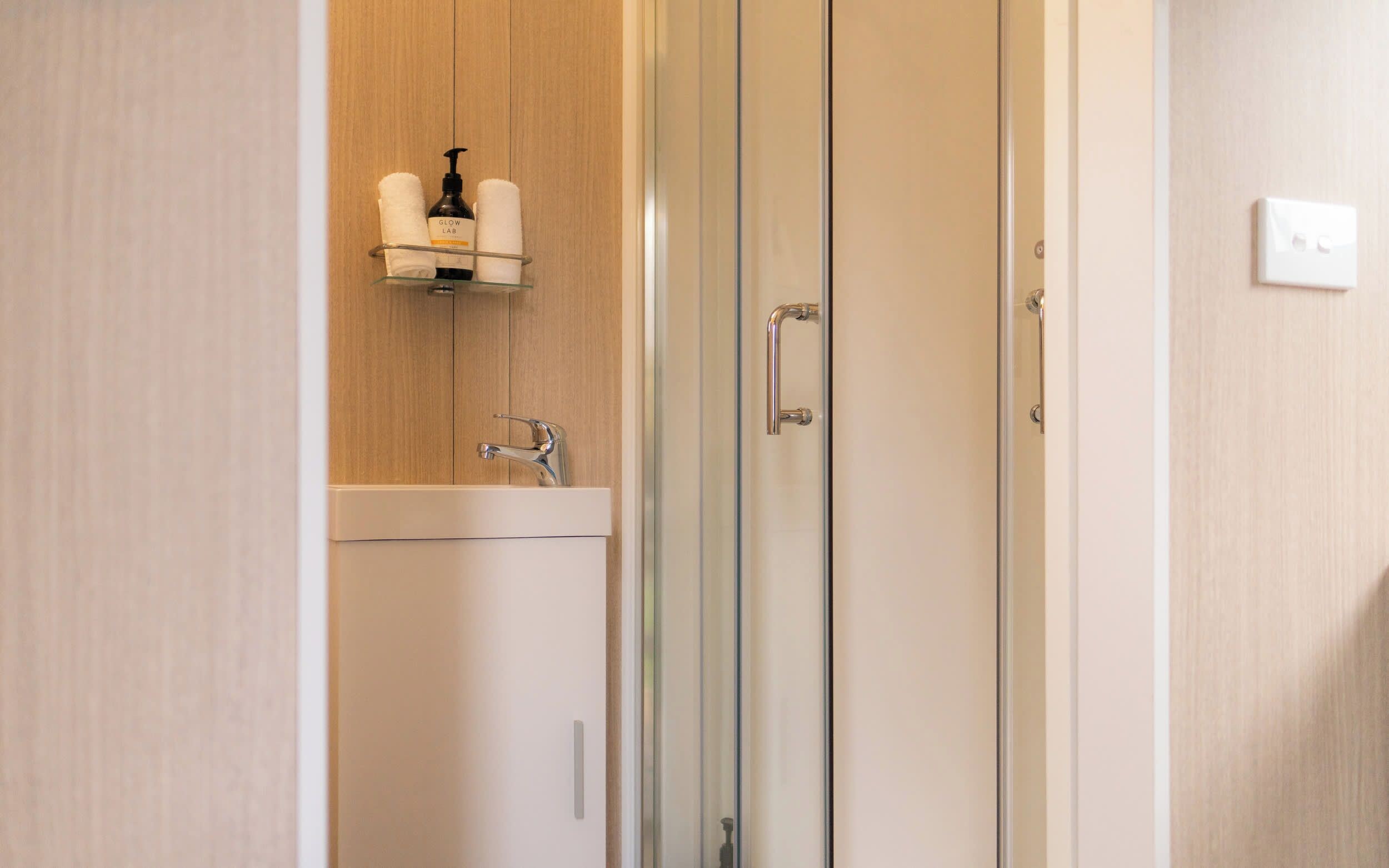 A modern bathroom with a glass shower door, a white sink, and a towel rack with a bottle of shampoo.