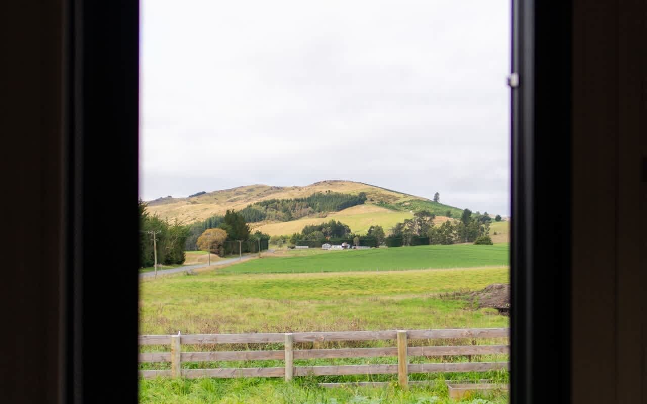Une vue à travers une fenêtre sur un paysage rural, avec une colline, des champs verdoyants et une clôture en bois.