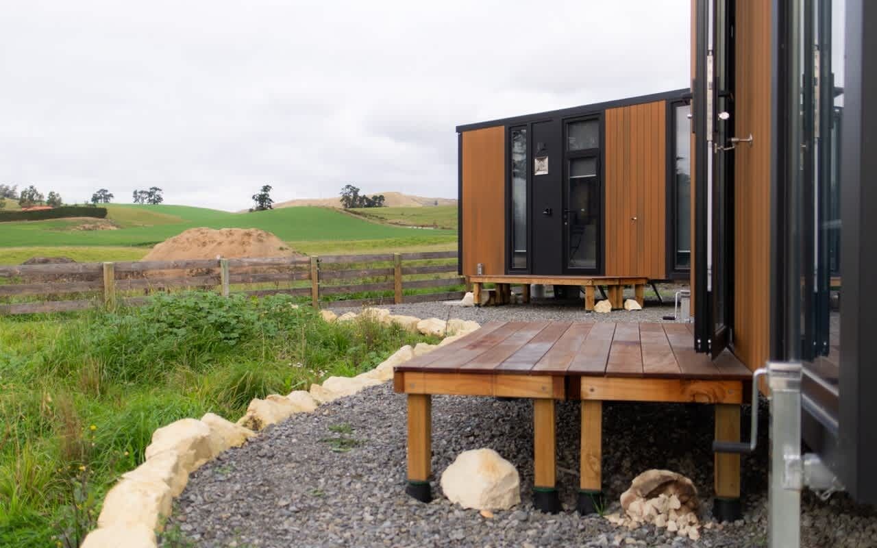 A modern cabin with a wooden deck, situated in a rural landscape with rolling hills and a fenced field.