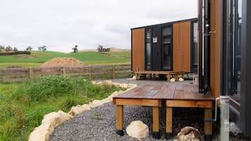 A modern cabin with a wooden deck, situated in a rural landscape with rolling hills and a fenced field.
