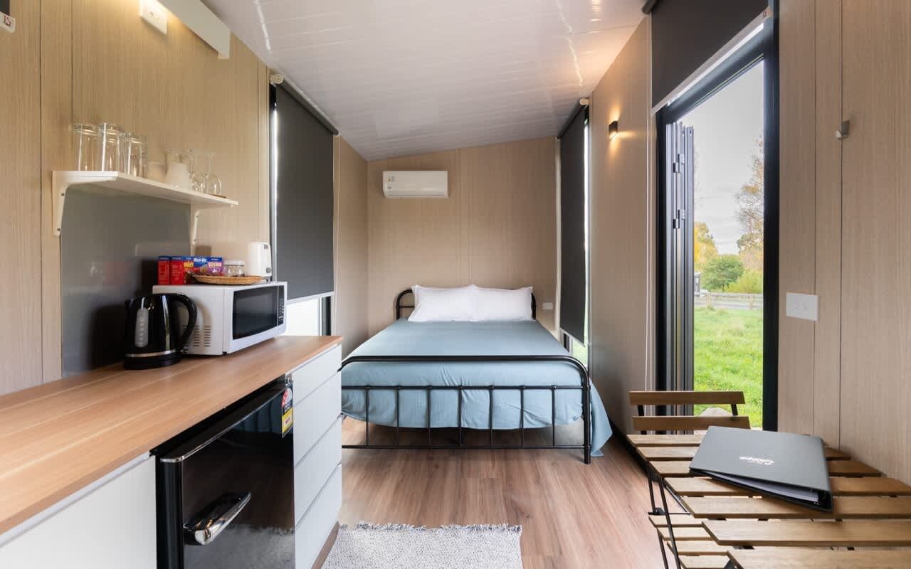 Habitación pequeña con cama, kitchenette, ventana y un ordenador portátil sobre una mesa.