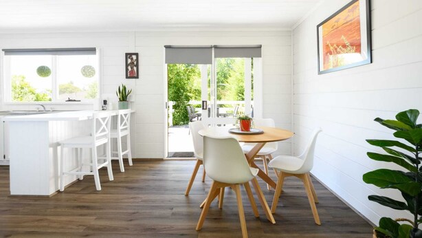 A bright kitchen with white cabinetry, a wooden dining table with white chairs, and a view of the outdoors through large windows. - Savanna Lodge Daylesford (Daylesford)
