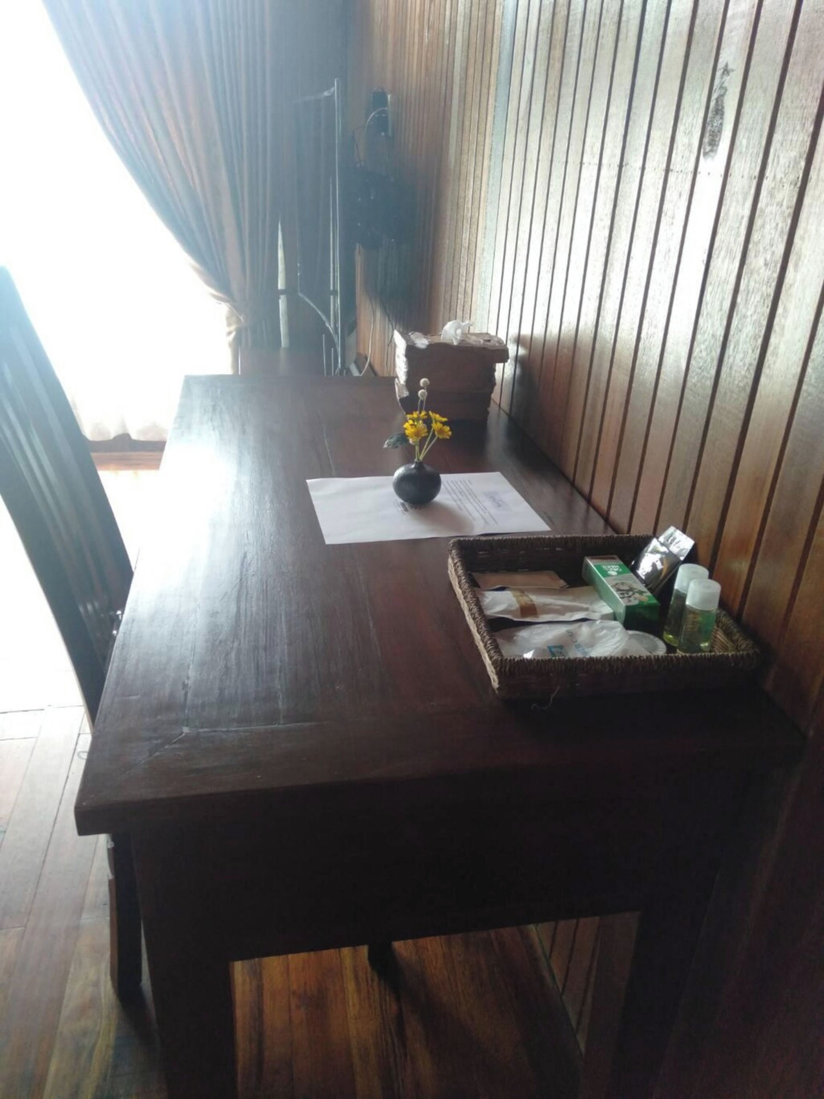 A wooden table with a vase of yellow flowers, a tray with toiletries, and a piece of paper on it.