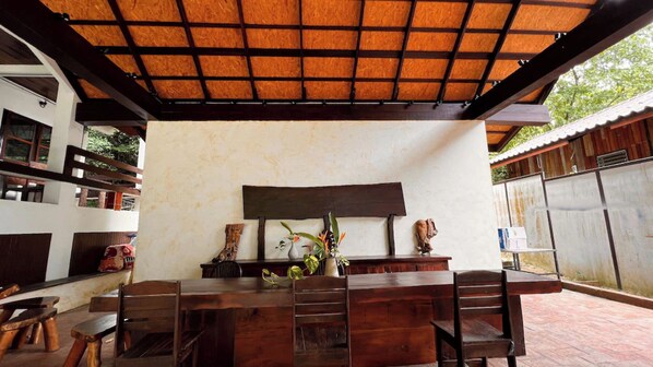 Un espace bar extérieur avec une table et des chaises en bois, un grand bac à plantes, et une clôture en métal en arrière-plan.