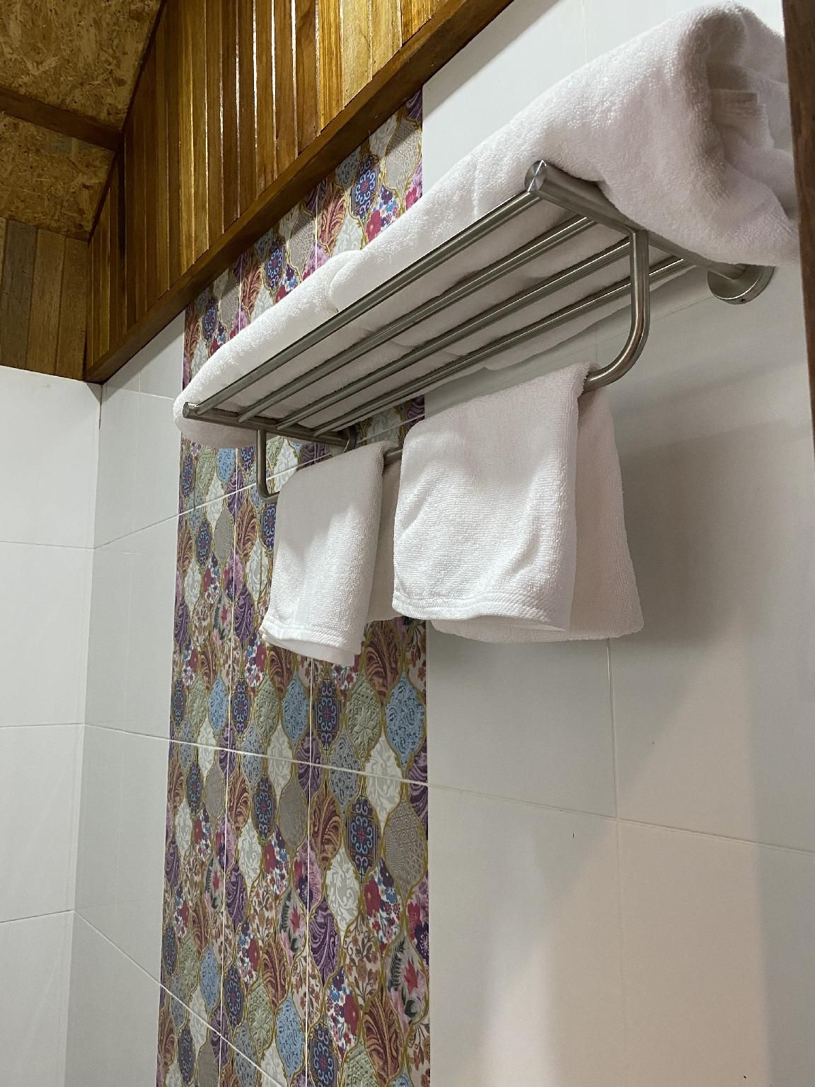 A bathroom with a metal towel rack holding two white towels, a tiled wall with a floral pattern, and a wooden ceiling.