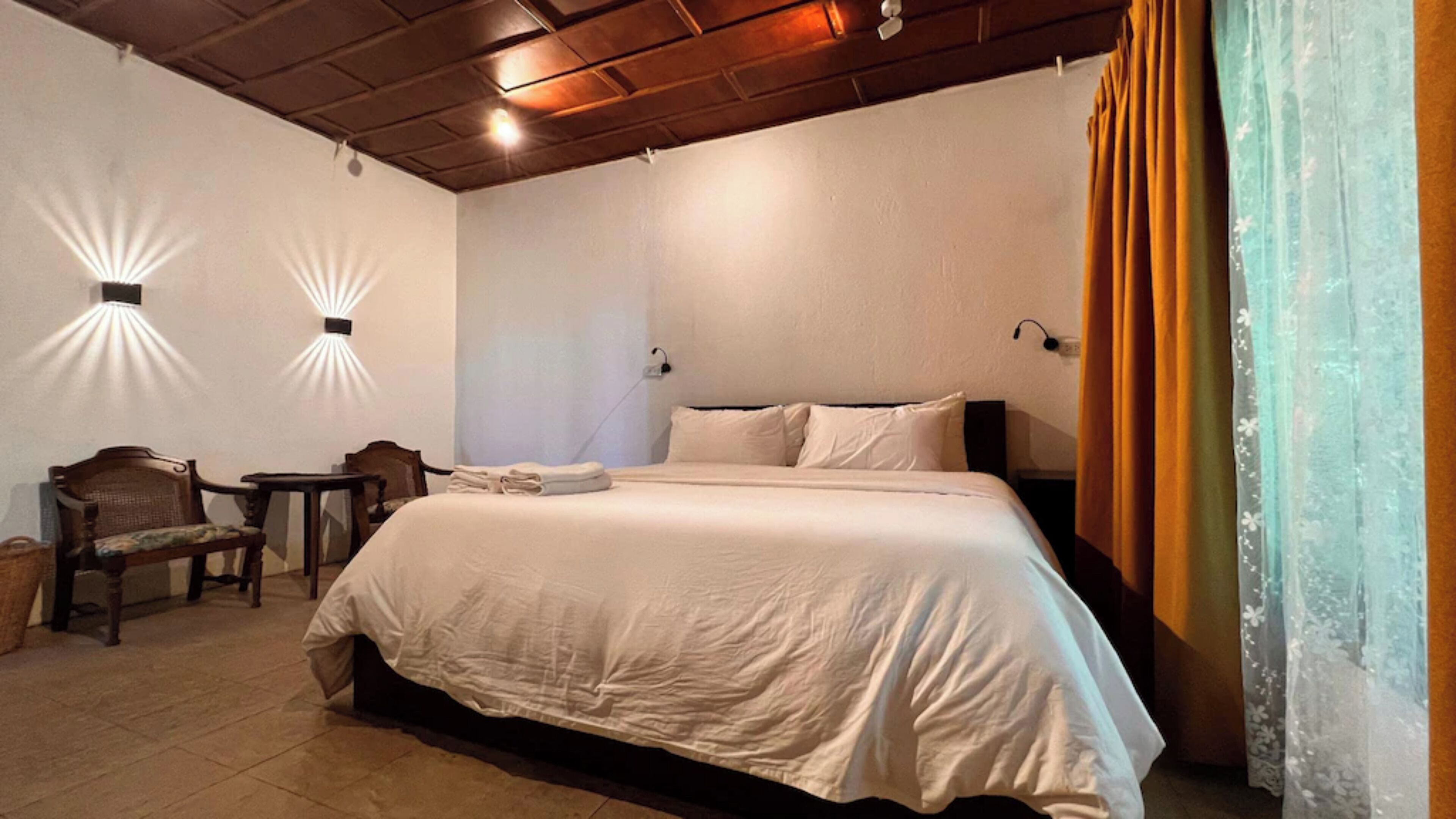 A bedroom with a large bed, two wall-mounted lights, a chair, and a small table.