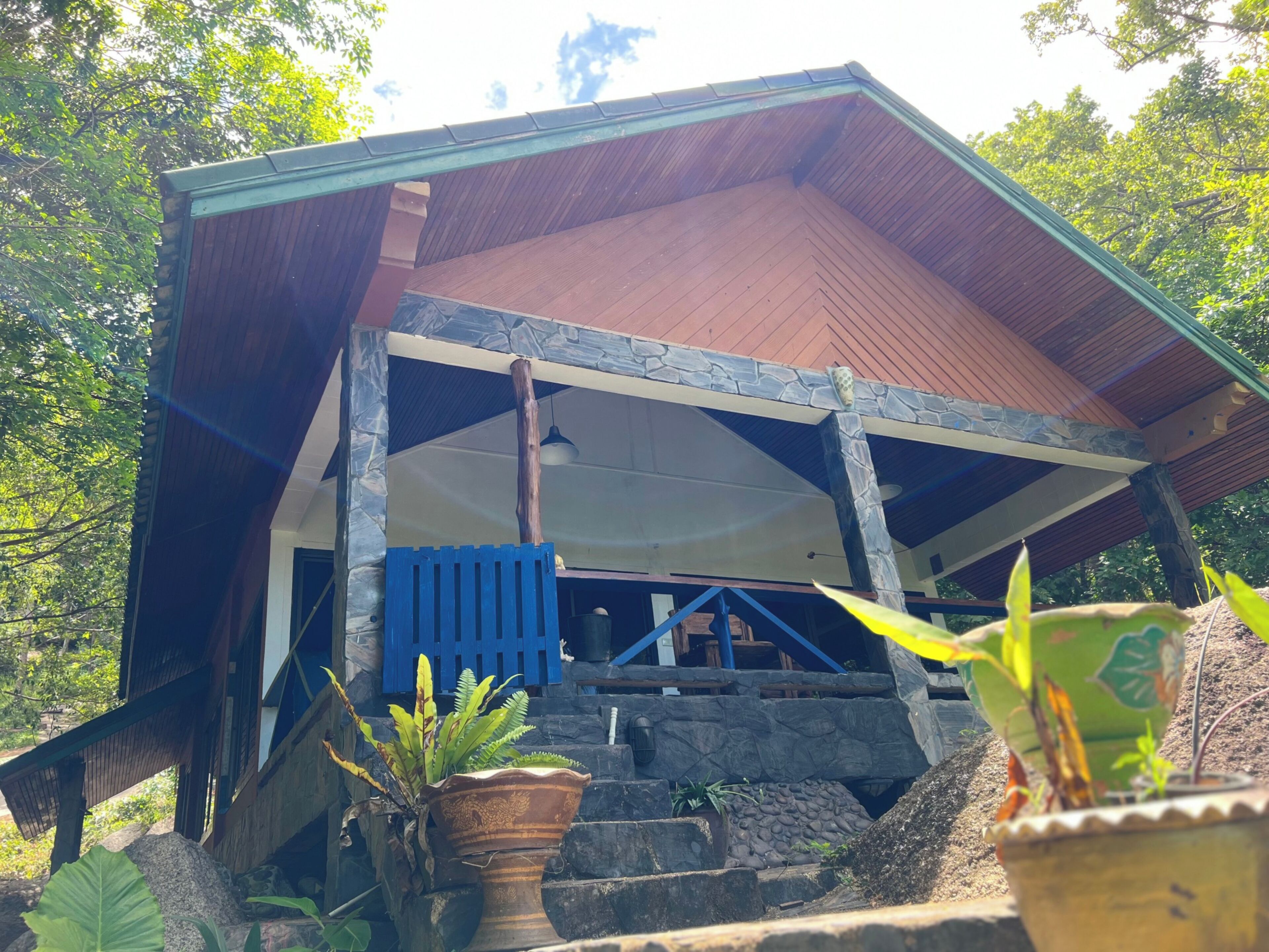 Un edificio rústico con fachada de madera y cimientos de piedra. Tiene un porche con barandilla azul y una escalera de piedra que conduce a él.