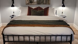 A neatly made bed with a metal frame, two bedside tables with lamps, a large painting of cacti on the wall, and a patterned rug on the floor. - 4bd 3ba - Pool / HotTub/ Arcade/ Theater/ PingPong/ Foosball (Pflugerville)