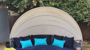 A curved outdoor seating area with a canopy, featuring a dark blue cushioned bench with bright blue cushions, a wicker table, and a beverage tray with drinks. - 4bd 3ba - Pool / HotTub/ Arcade/ Theater/ PingPong/ Foosball (Pflugerville)