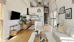 A spacious living area with a sofa, a coffee table, and a TV. There is a kitchen area with a stove, sink, and cabinets. A staircase is visible in the background.