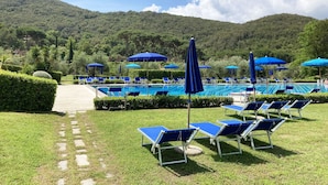 Zona de piscina con sillas reclinables azules y sombrillas, rodeada de césped y setos cuidados, con montañas al fondo.
