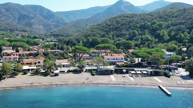 Un pueblo costero con edificios a lo largo de la playa, un muelle que se adentra en el agua y montañas al fondo.