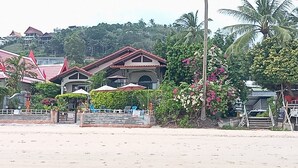 A beachfront property with multiple buildings, some with red roofs, and lush greenery including palm trees and flowering plants.