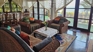 A spacious living room with wicker furniture, a dining area with wooden chairs, and large windows with curtains.
