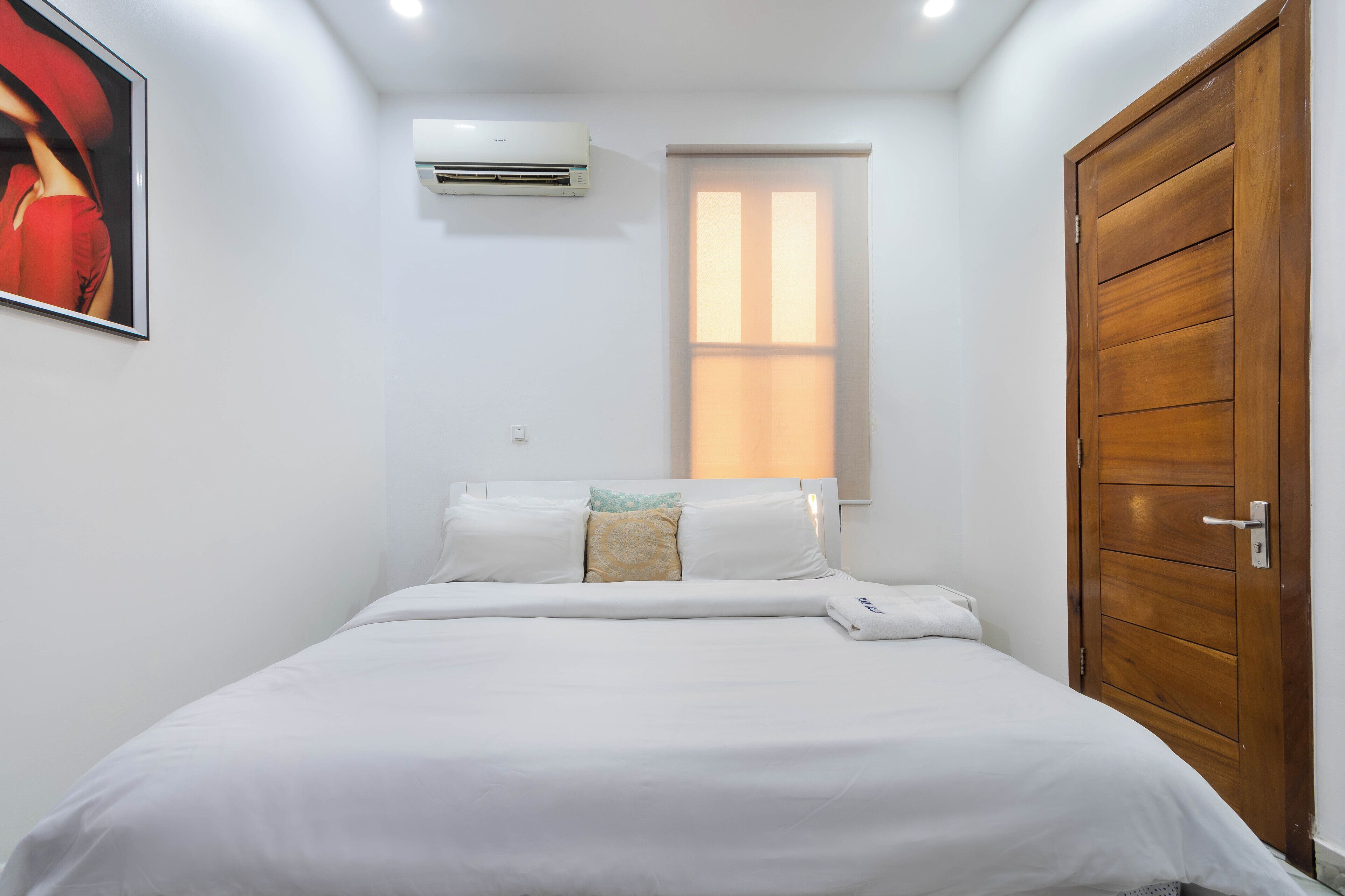 A bedroom with a bed, white bedding, a wooden door, a window with blinds, and an air conditioning unit on the wall.