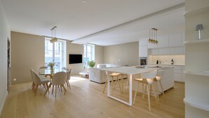 Una cocina moderna con una isla central, un comedor con mesa de vidrio y una sala de estar con sofá y televisor.