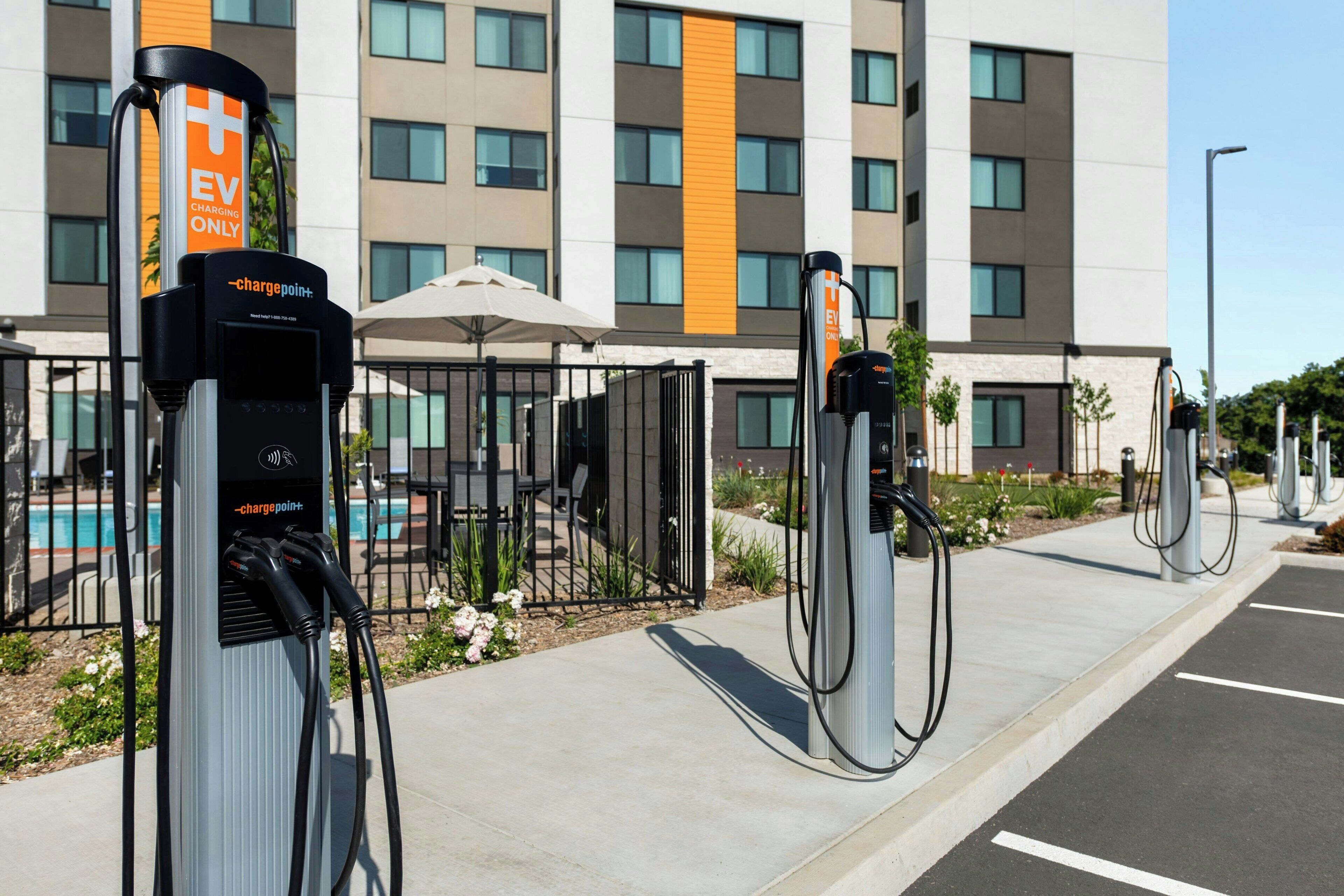 A row of EV charging stations in front of a modern building with a pool and outdoor seating.