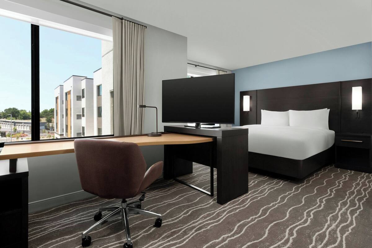 Een hotelkamer met een bureau, een groot flatscreen televisietoestel, een bed met witte lakens, een bruine bureaustoel en uitzicht op gebouwen door het raam.