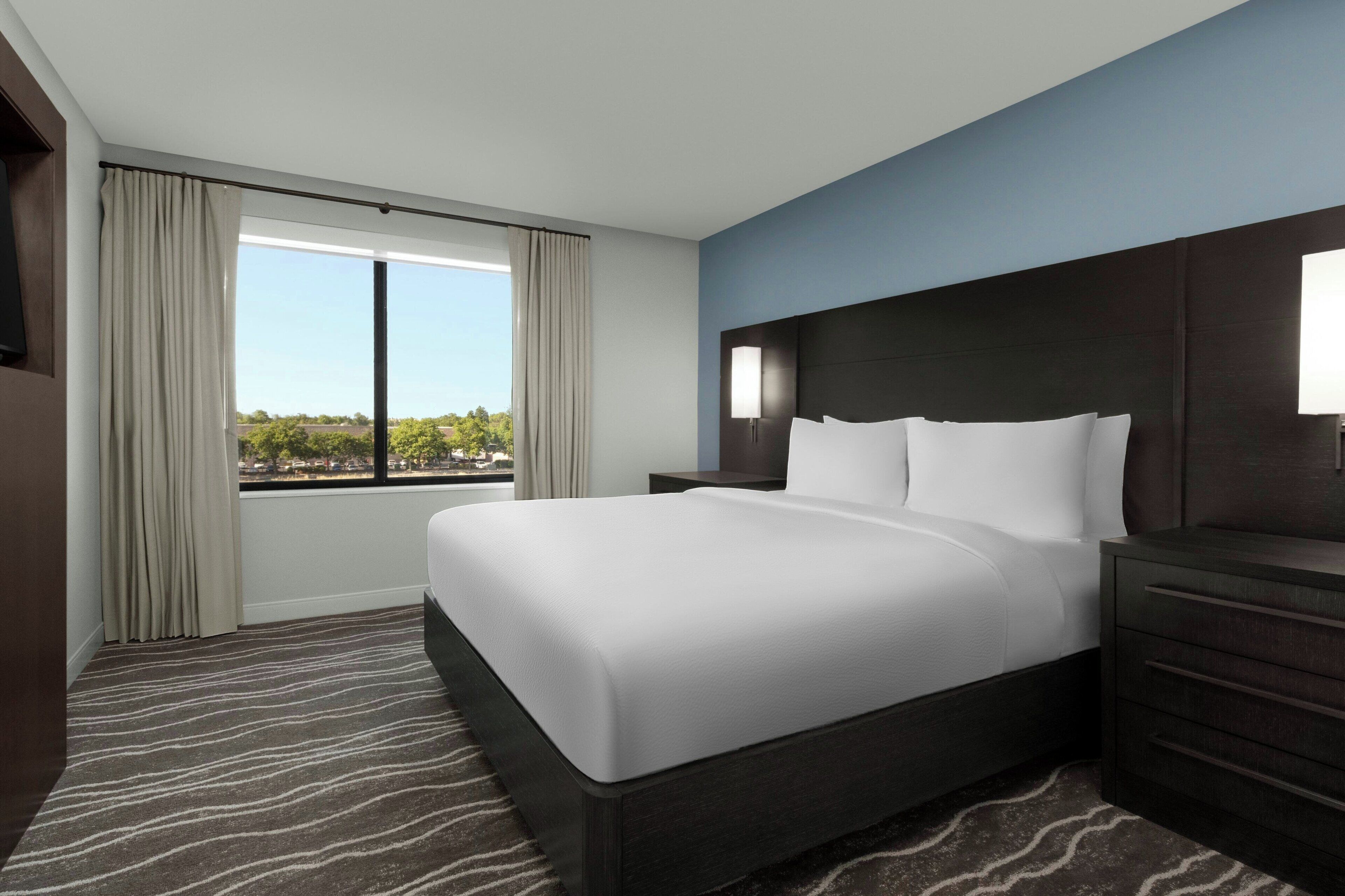 A hotel room with a large bed, a nightstand, a dresser, and a window with curtains.