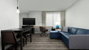 Habitación de hotel moderna con televisor de pantalla plana, escritorio con silla, sofá azul y alfombra estampada.