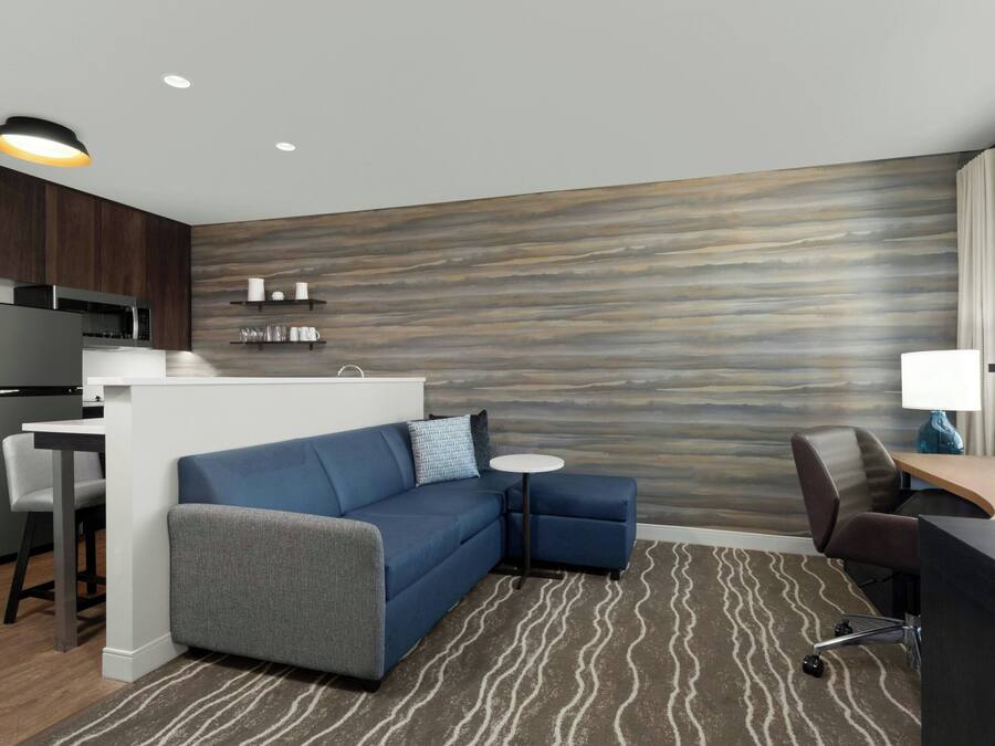 A modern hotel room with a kitchenette, a blue sofa, a small round table, and a desk with a lamp.