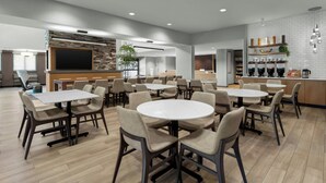 A modern dining area with multiple tables and chairs, a kitchen area with a bar, and a flat-screen TV mounted on a brick wall. - Residence Inn by Marriott Sacramento Cal Expo (Sacramento)