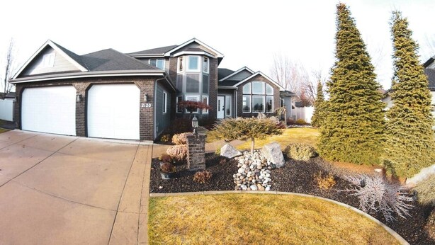 A two-story house with a garage, a well-maintained lawn, and two tall evergreen trees. - Gorgeous Executive Home - Hot Tub, Gym, Theatre, and More! (Spokane)